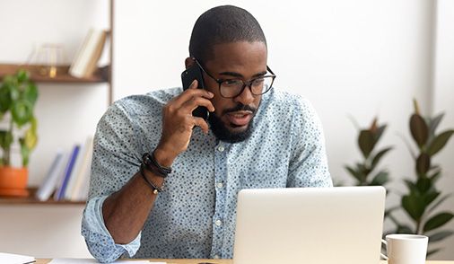 Man pictured sitting at modern in-home office desk on laptop, speaking on his mobile phone