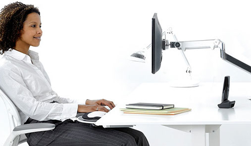 Working woman smiling while sitting at desk, set up with adjustable solutions on her computer monitor, arm, and foot rests.